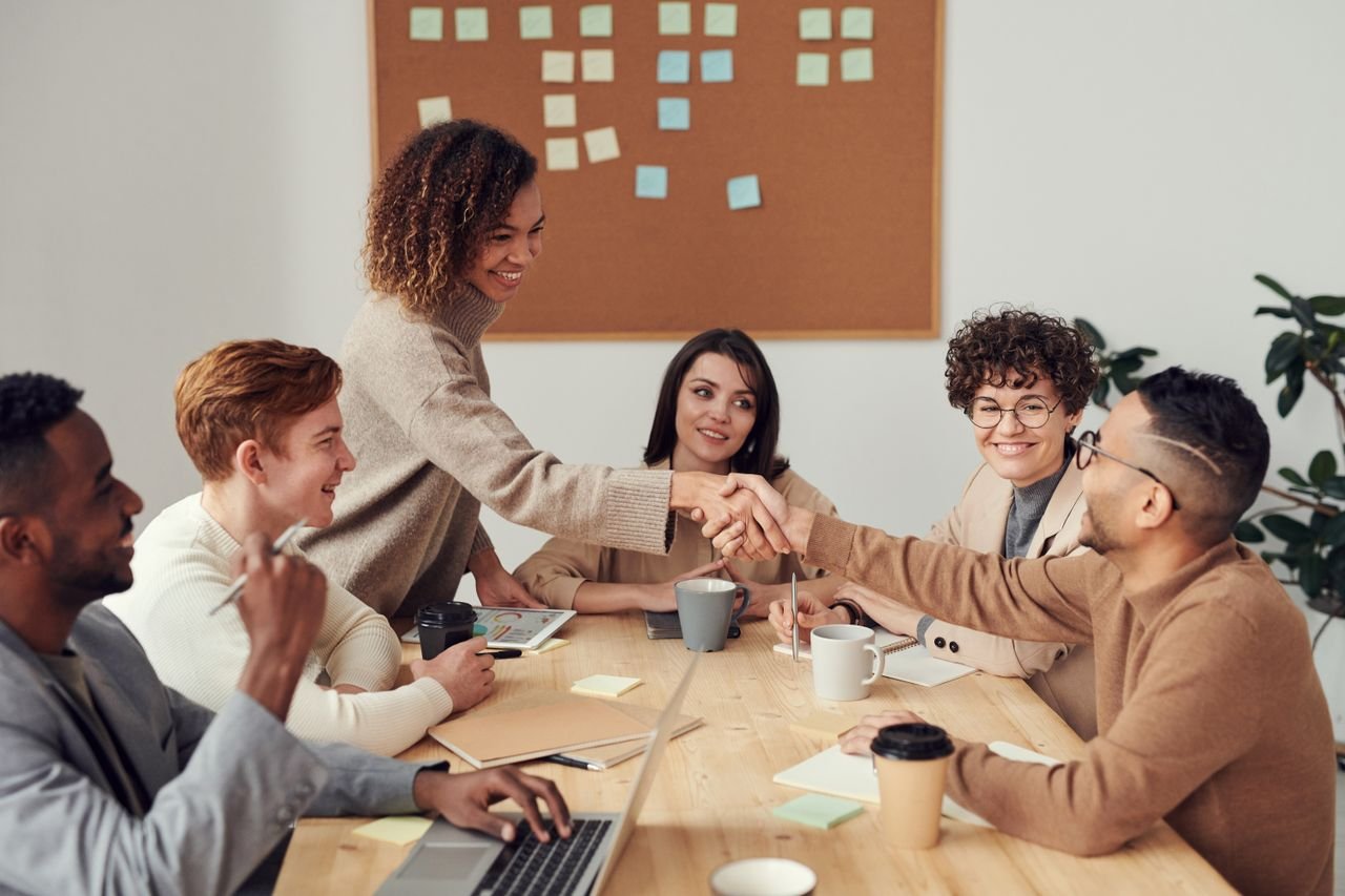Team members shaking hands across a meeting table with laptops and coffee